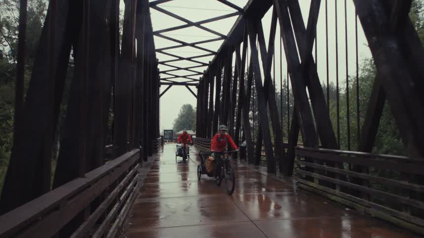 A pair of determined cyclists in red raincoats pull trailers across a wet, wooden truss bridge on a rainy day