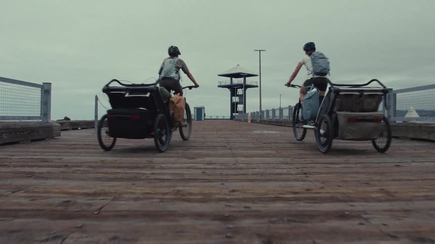 A low angle shot follows two people as they ride their bicycles with gear trailers down a long wooden pier on a cloudy day