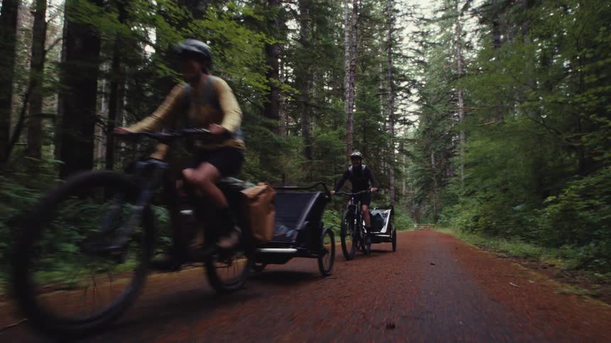 A slow-motion tracking shot follows two adventurous cyclists with trailers as they ride along a leaf-strewn path in a lush forest