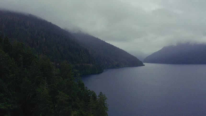Moody aerial footage captures the serene beauty of a vast lake and forested mountains shrouded in fog and clouds