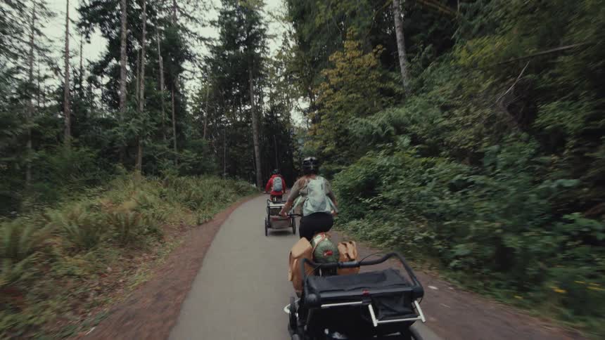 A point-of-view shot follows two cyclists with trailers as they bike-pack on a paved trail through a dense evergreen forest