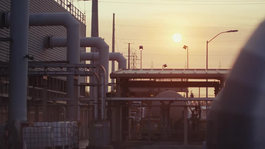 A slow dolly shot reveals an industrial factory with extensive pipework against a glowing sunset
