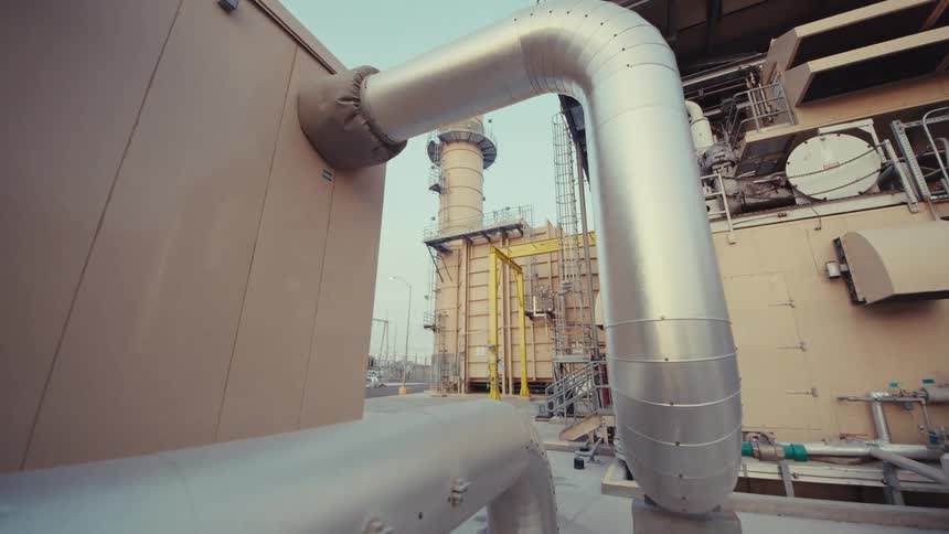 A slow dolly shot moves through the complex infrastructure of a modern power plant, revealing its intricate network of pipes and machinery