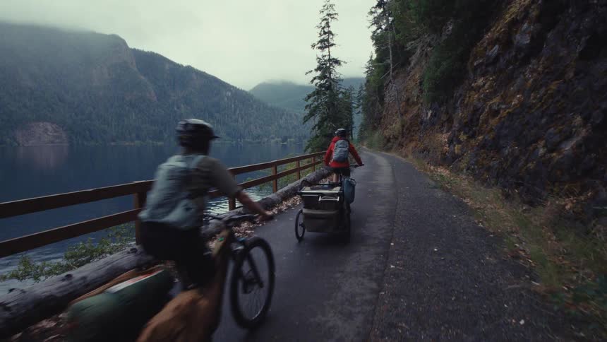 A following shot of two cyclists on a bike-packing adventure as they pedal along a scenic lakeside path in the mountains