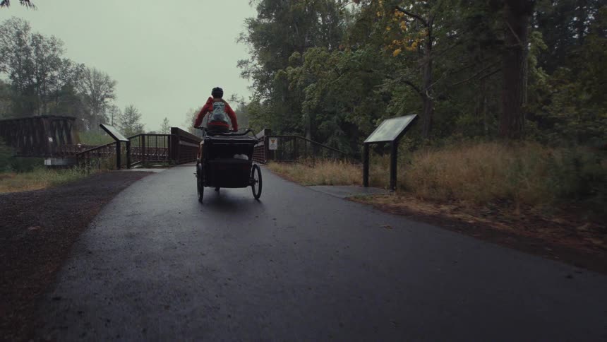 A pair of cyclists in red jackets tow trailers on a bike tour along a wet, paved trail through a wooded park
