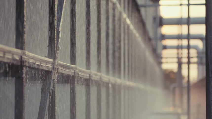 Slow motion close up of a water cooling system spraying water down a long industrial wall at sunset