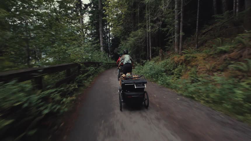 A dynamic tracking shot follows two bikers pulling gear trailers on a scenic trail through a lush forest
