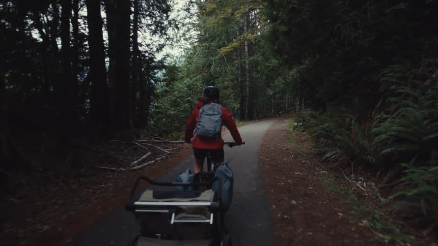 A couple on an adventurous bikepacking trip ride their bikes with trailers down a paved path through a dense, green forest