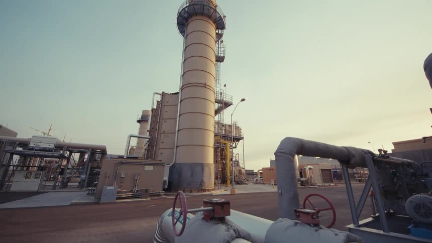 A slow reveal of an industrial power plant, starting from the intricate network of pipes and moving towards the towering smokestacks at sunset