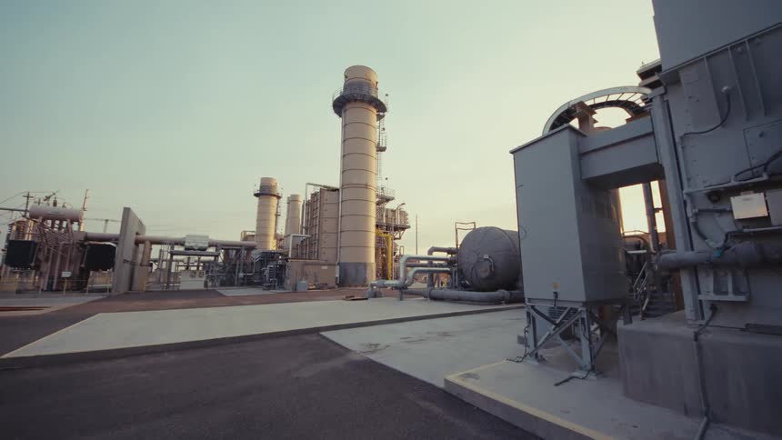 A slow dolly shot moves through the complex infrastructure of a power generation facility as the sun rises