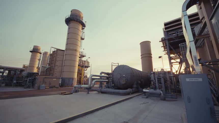A slow push-in shot reveals an expansive industrial power plant with large towers, pipes, and machinery at sunset