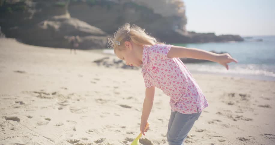 In slow motion, a little girl with a shovel plays on a sunny beach before running towards the gentle waves