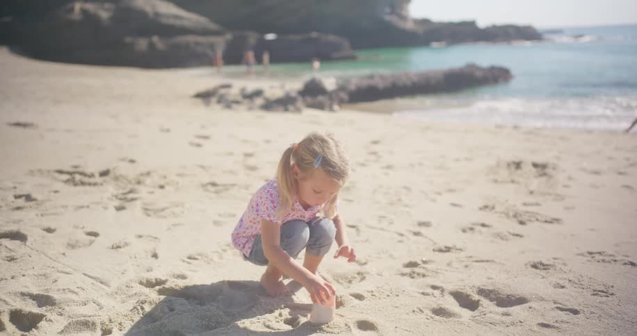 A young girl with blonde pigtails carefully sets down a cup of sand while playing on a sunny beach