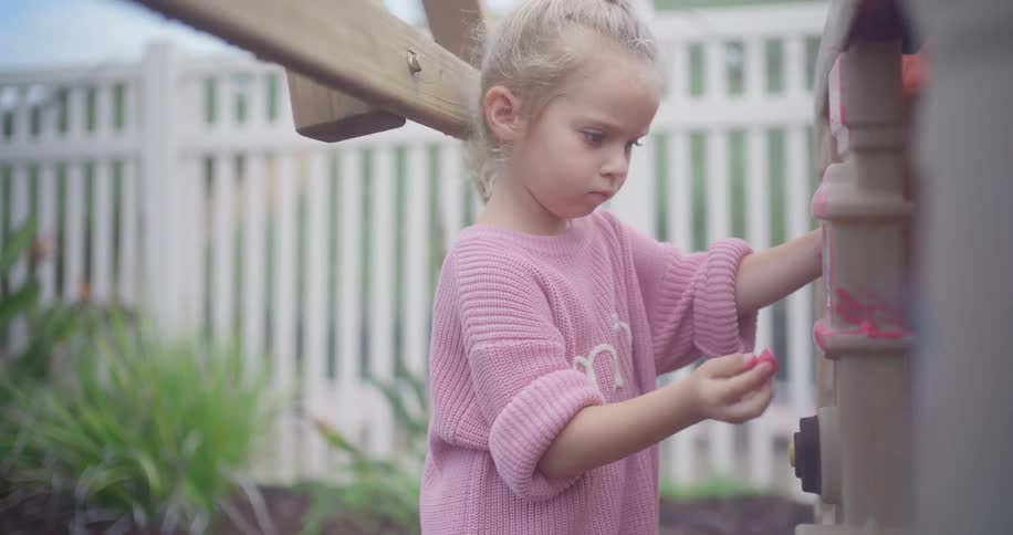 A young girl in a pink sweater concentrates while playing with a plastic climbing wall in her backyard