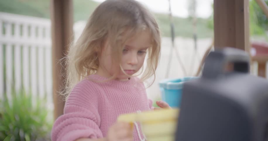 Intensely focused little girl in a pink sweater carefully pours water from a yellow cup in slow motion