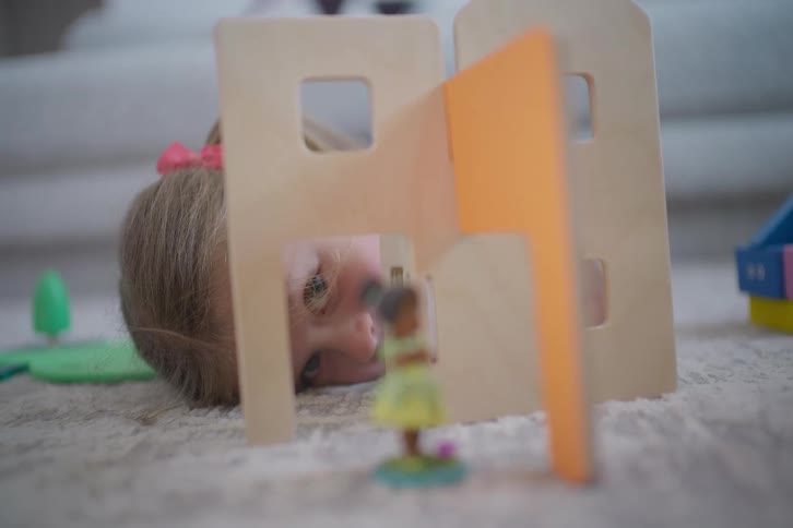 A little girl's eye peeks inquisitively through the window of a wooden toy dollhouse on the floor