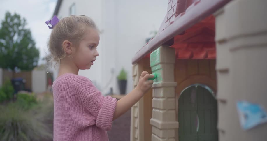 A young girl in a pink sweater happily colors on her plastic playhouse with green chalk in the backyard