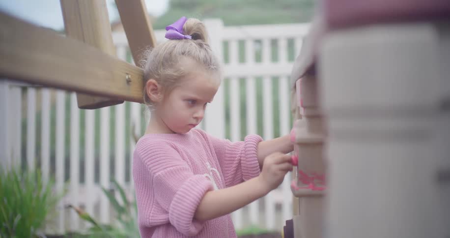 A sweet young girl with a blonde ponytail and a pink sweater plays intently on her backyard playset