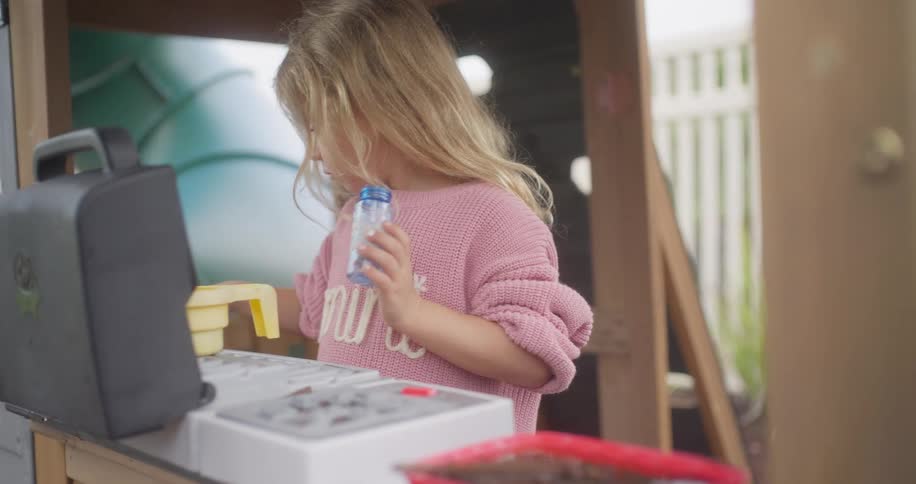 A young blonde girl in a pink sweater plays chef with her toy kitchen set in a backyard playhouse