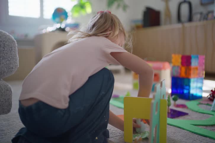 A focused young girl with blonde hair kneels on the carpet while playing with her colorful toy set in a bright living room