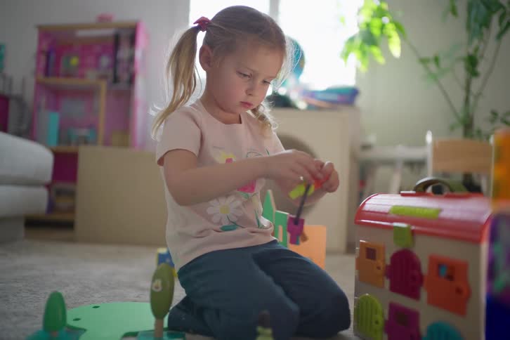 A focused little girl kneels on the floor in her playroom, carefully trying different keys to unlock a colorful educational toy