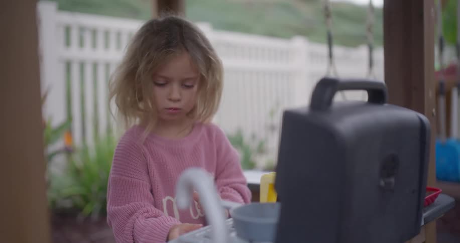 A cute little girl in a pink sweater concentrates while playing with her toy kitchen and grill in the backyard
