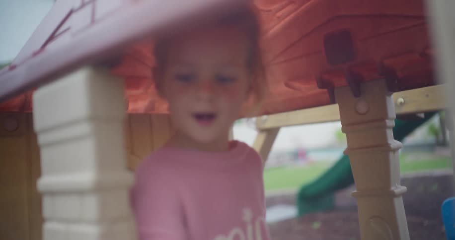A young girl with a purple bow plays with stickers in her playhouse at the park, looking up happily at the camera