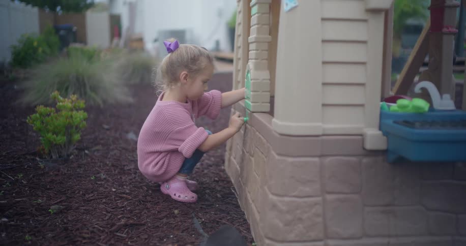 A cute little girl with a purple bow in her hair crouches down to carefully color on her plastic playhouse in the backyard