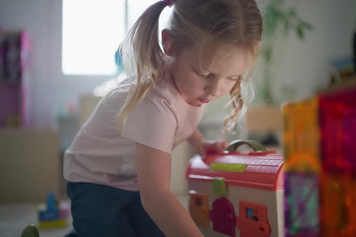 Little girl plays with a colorful toy house on the floor in a sunlit playroom