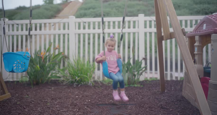 Little girl in pink sweater gently swinging on backyard playset near white fence