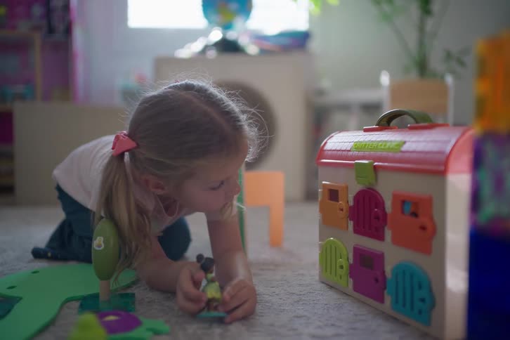 A little girl with pigtails kneels on the floor, deeply engaged in playing with her colorful toys in a sunlit room