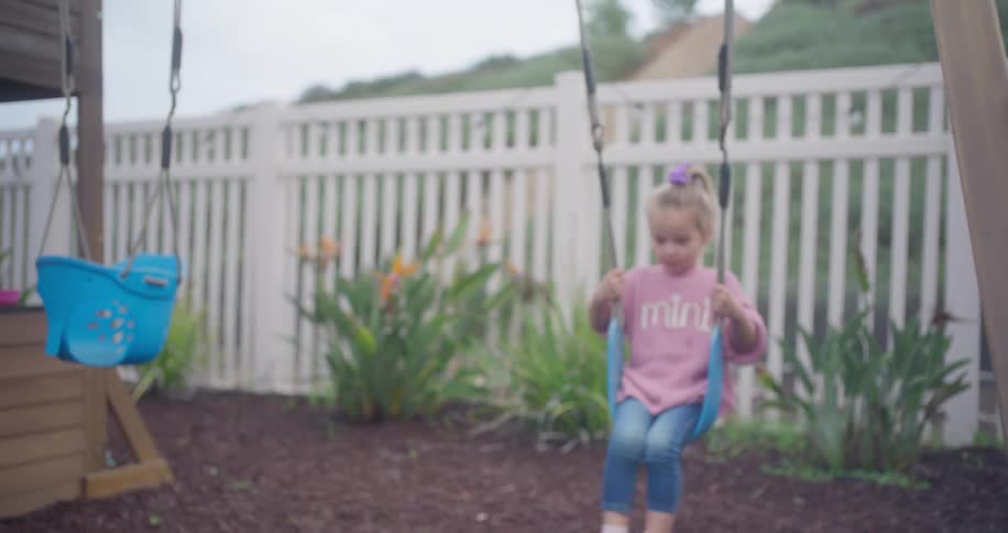 A cute little girl in a pink sweater happily swings back and forth on a playground in her backyard in slow motion