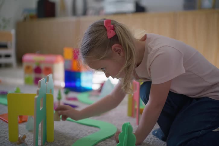 A focused young girl with a pink bow in her hair kneels on the carpet and plays with a colorful toy village set in a sunlit room