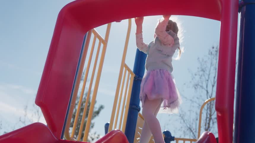 Happy toddler girl in a tutu hangs from a colorful playground bar in slow motion as the bright sun shines