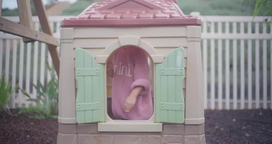 An adorable little girl in a pink sweater plays in her backyard playhouse, holding up a treat and smiling from the window