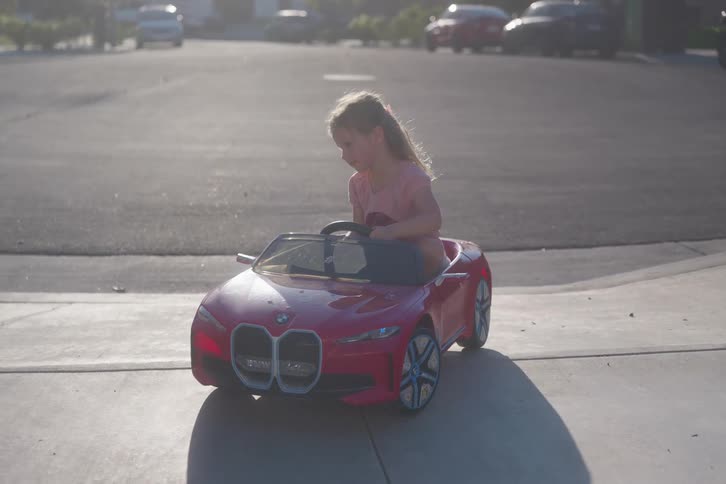 A cute little girl drives her red toy convertible car down the sidewalk in slow motion on a sunny day