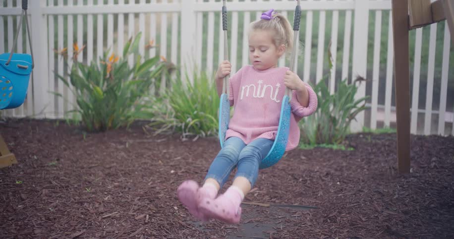 A cute little girl with a ponytail gently swings back and forth on a playset in a backyard, filmed in slow motion