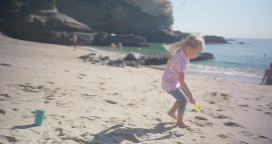 In slow motion, a young girl with pigtails plays on a sunny beach, filling a cup with sand from a small shovel