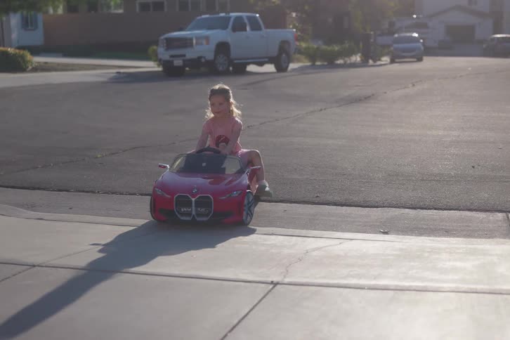 A young girl sits in her red toy convertible car at the edge of a driveway in a quiet suburban neighborhood during golden hour