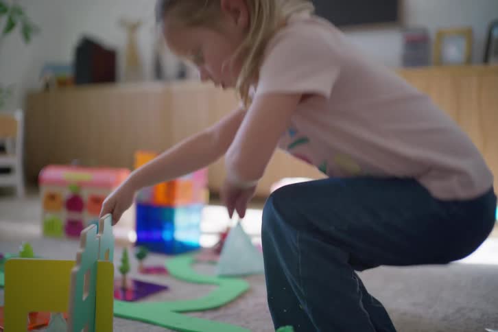 A young blonde girl in slow motion plays with colorful building blocks on the carpet of her living room