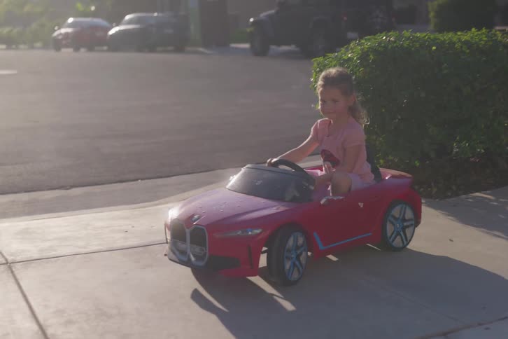 An adorable little girl happily drives her red convertible toy car down the sidewalk in a sun-drenched neighborhood in slow motion