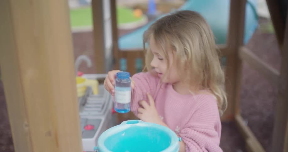 Adorable little girl in a pink sweater uses her imagination to cook with a toy kitchen set at an outdoor playground