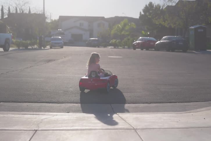 A joyful little girl drives her red convertible toy car down a suburban cul-de-sac in slow motion on a sunny afternoon