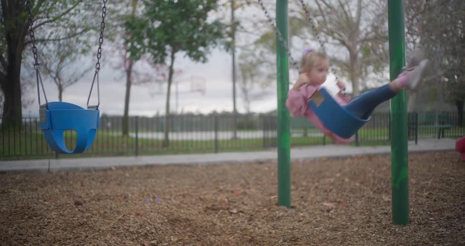 A cute little girl in a pink sweater gently swings back and forth in slow motion on a playground swing set
