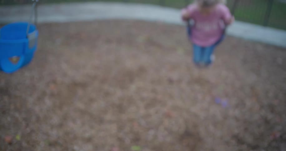 A young girl with a pensive expression swings gently back and forth in slow motion on a playground swing set