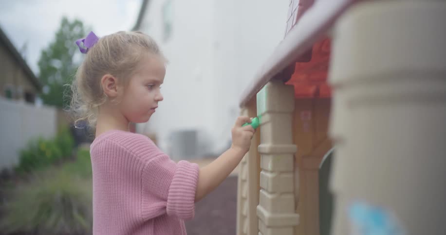 A focused little girl with a purple bow in her hair carefully draws with green chalk on her plastic playhouse in the backyard
