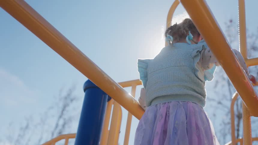 Slow motion from below of a little girl in a tutu climbing across yellow monkey bars on a sunny day