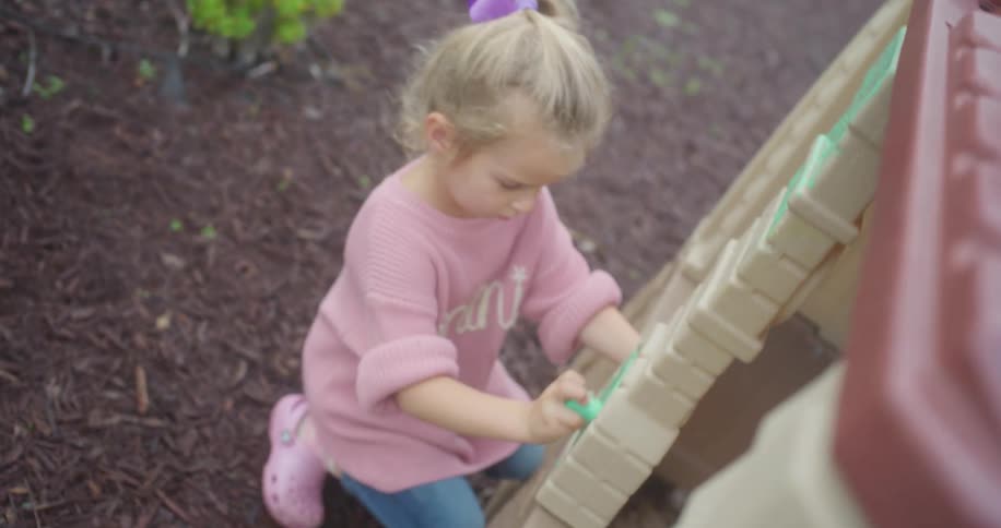 High-angle slow-motion shot of a little girl in a pink sweater drawing with green chalk on her backyard playset