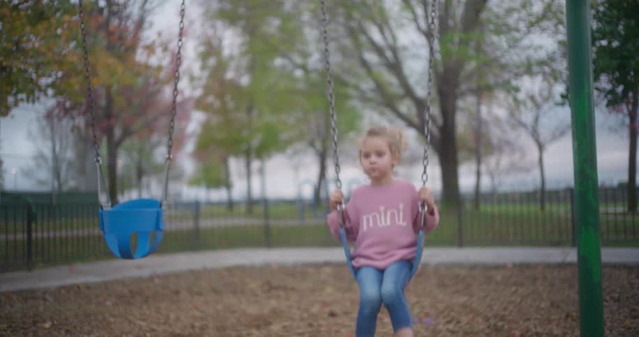 A little girl in a pink sweater enjoys swinging on a playground swing set as the camera pulls back in slow motion