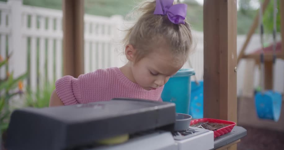 An adorable toddler girl in a pink sweater and purple bow smiles as she plays pretend with her toy kitchen in the backyard in slow motion